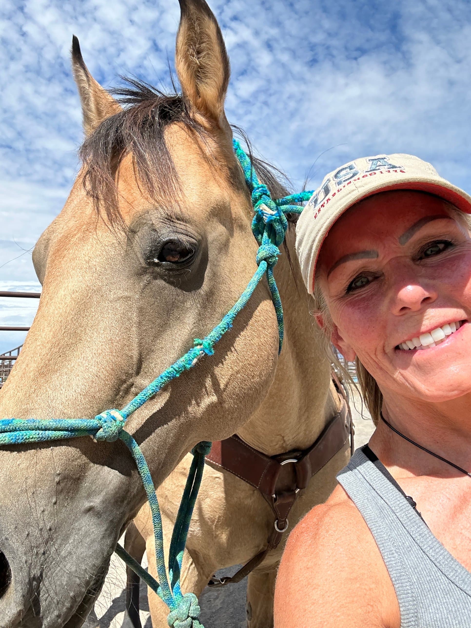 Mary Leeds in a USA cap taking a selfie beside her buckskin quarter horse under a blue sky