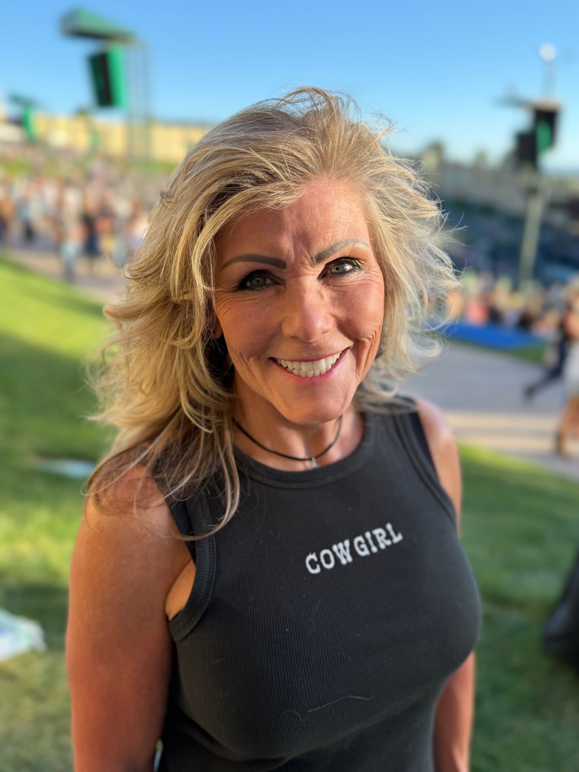 Mary Leeds smiling outdoors in a black tank top printed with the word COWGIRL, with a sunny event lawn behind her